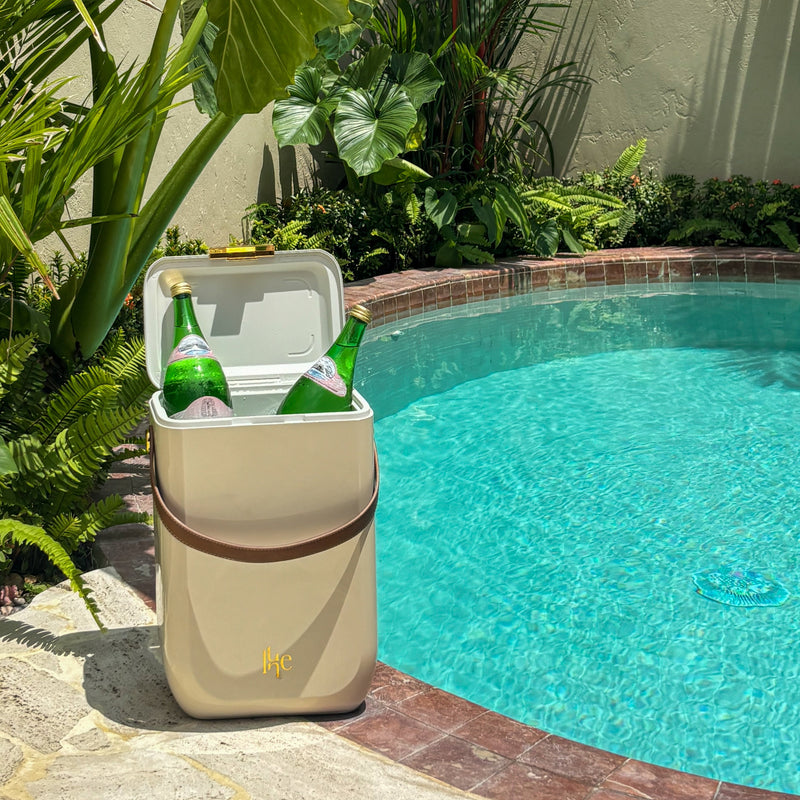 A beige insulated cooler bag with a brown handle sits beside a swimming pool, with green bottles inside and tropical plants in the background.